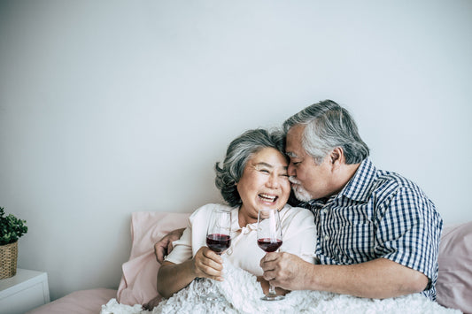 Elderly couple laying in bed holding red wine glasses. 