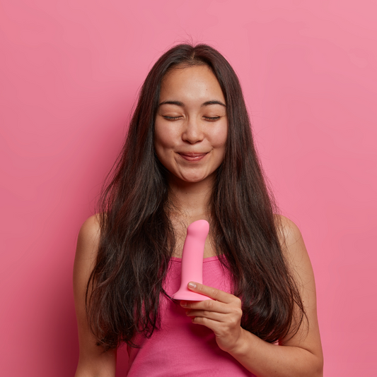 Woman smiling while holding a pink dildo while standing in front of a pink background. 