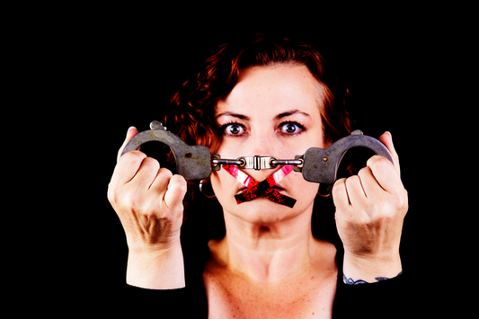 A dark red-haired woman is holding up handcuffs while her mouth is taped shut.